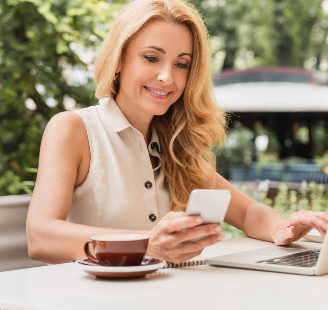 Vertical photo of successful mature businesswoman working remotely on distance on laptop, using smart phone in cafe. E-banking, e-commerce concept